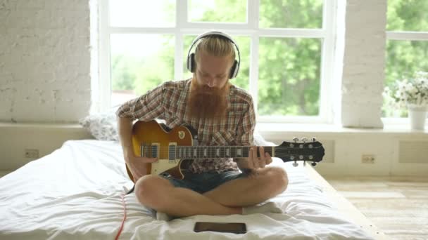 Homem barbudo atraente em fones de ouvido sentado na cama aprendendo a tocar guitarra usando o computador tablet no quarto moderno — Vídeo de Stock