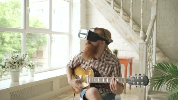 Ung bearded man sittande på stol lära sig spela gitarr med hjälp av Vr 360 headset och känns honom gitarrist på konsert hemma — Stockvideo