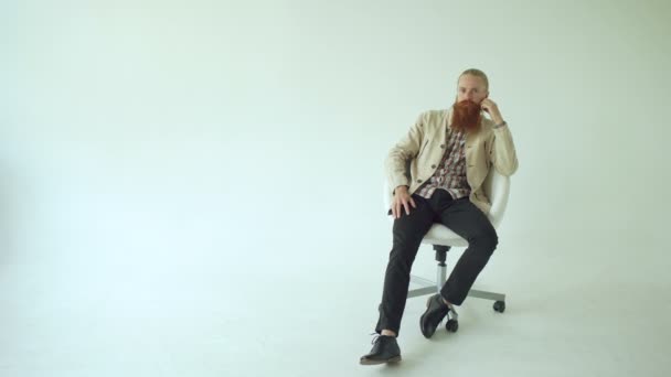 Серйозний бородатий чоловік сидить на поворотному офісному стільці і дивиться в камеру на білому тлі — стокове відео