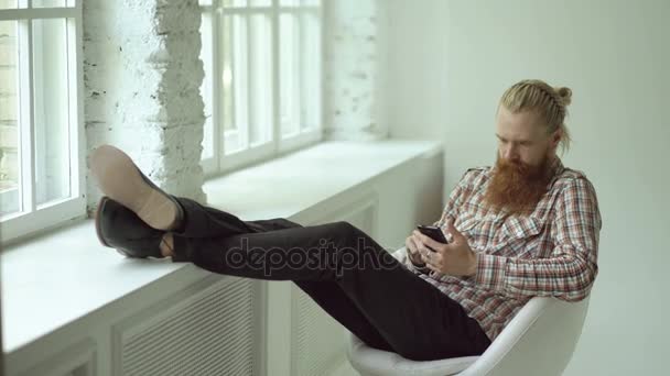 Skäggig ung hipster man sms-textning med smartphone medan du sitter i offce stol med sina ben på fönsterbrädan — Stockvideo