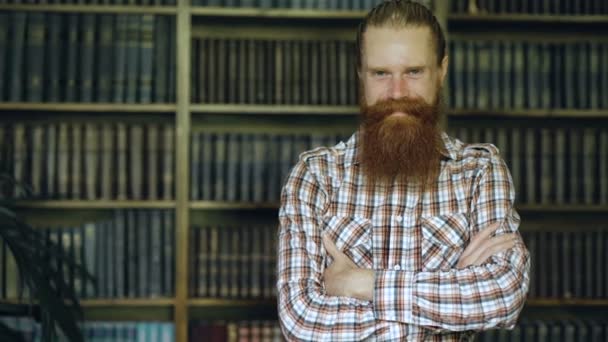 Portret jonge bebaarde man die lacht gelukkig in de bibliotheek en op zoek naar camera — Stockvideo