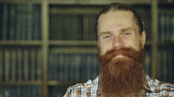 Портрет крупным планом молодой бородатый мужчина улыбается счастливо в библиотеке и смотрит в камеру — стоковое видео