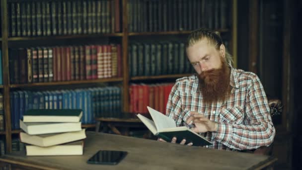 Joven estudiante barbudo en la biblioteca leyendo un libro y preparándose para los exámenes — Vídeo de stock