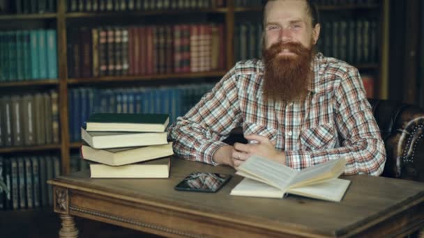 Omhoog kantelen van portret jonge bebaarde man die lacht gelukkig in de bibliotheek en op zoek naar camera — Stockvideo