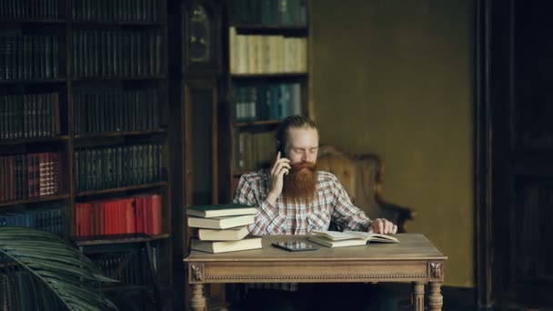 Jovem barbudo estudante falando telefone enquanto se prepara para exames na biblioteca da Universidade — Vídeo de Stock