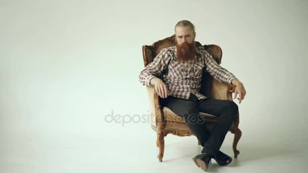 Bebaarde man zitten Luxe fauteuil en op zoek naar camera op witte achtergrond — Stockvideo