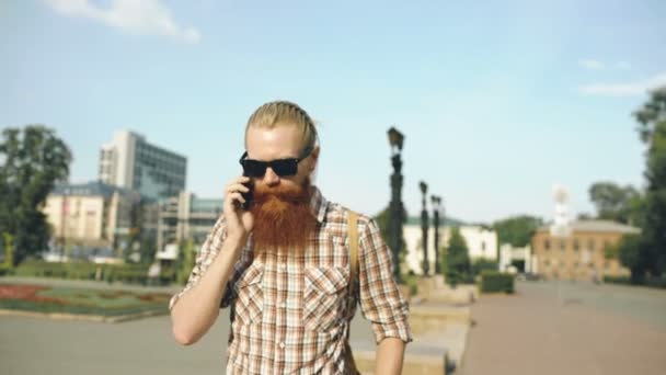 Steadicam снимок улыбающегося мужчины с рюкзаком, идущего по улице и говорящего по телефону на улице — стоковое видео