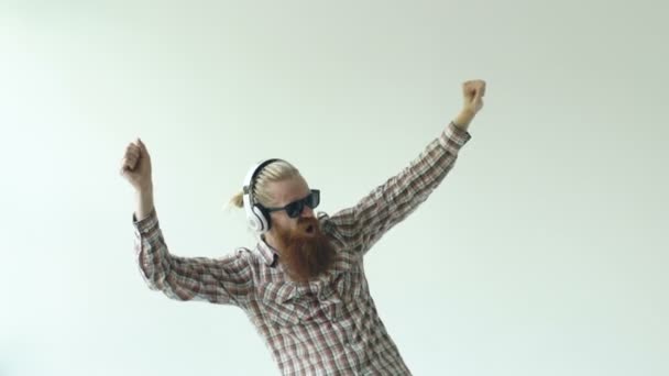 Счастливый молодой бородатый мужчина в солнечных очках и наушниках танцует и слушает музыку на белом фоне — стоковое видео