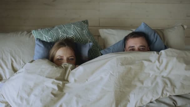 Vista superior de la pareja sonriente divirtiéndose en la cama escondida debajo de la manta y mirando a la cámara — Vídeo de stock