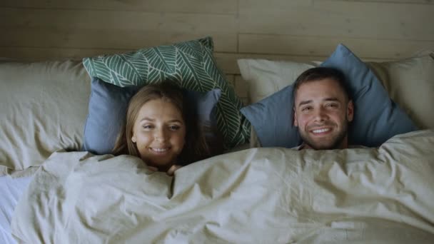 Top view of smiling couple having fun in bed hiding under blanket and looking into camera — Stock Video