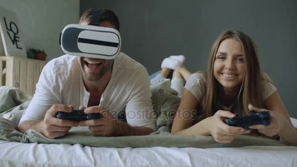 Casal jovem deitado na cama jogar jogos de vídeo com controlador e fone de ouvido VR — Vídeo de Stock