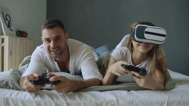 Pareja joven acostada en la cama jugar videojuegos con controlador y auriculares VR — Vídeo de stock