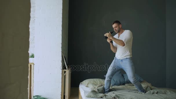 Young man singing to hair dryer and dancing on bed in bedroom — Stock Video