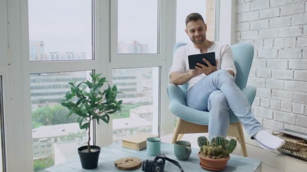 Aantrekkelijke man met behulp van digitale tablet zitten in de stoel op balkon in moderne loft-appartement — Stockvideo