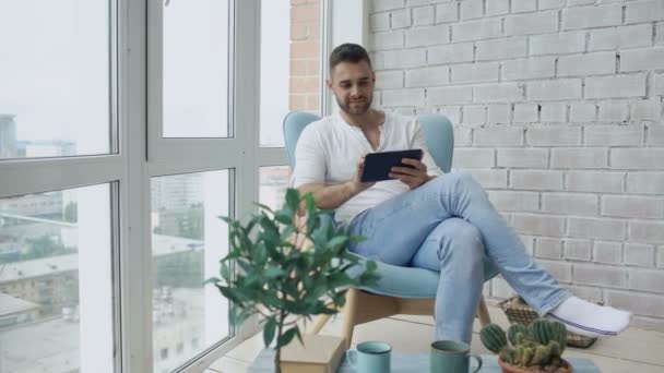 Atraktivní muž pomocí digitálních tabletu sedí v křesle na balkon, v podkroví moderní byt — Stock video