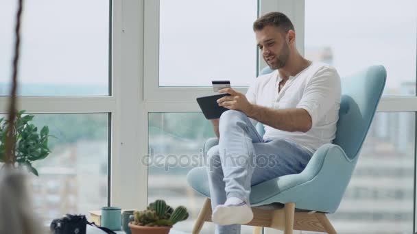 Молодой улыбчивый мужчина делает онлайн-покупки с помощью цифрового планшетного компьютера, сидя на балконе в современной мансардной квартире — стоковое видео