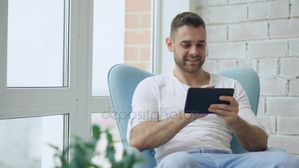 Улыбающийся мужчина с помощью цифрового планшета сидит в кресле на балконе в современной квартире — стоковое видео