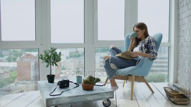 Молода приваблива жінка читає книгу і п'є каву, сидячи на балконі в сучасній квартирі лофт — стокове відео