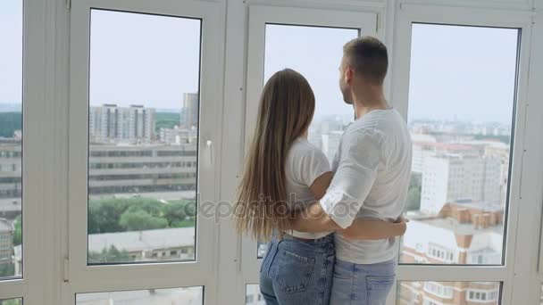 Joven pareja feliz abrazando de pie cerca de la ventana y disfrutando de la vista desde un nuevo apartamento — Vídeos de Stock