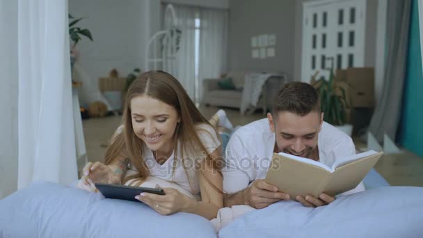 Νεαρό ζευγάρι έχετε ελεύθερο χρόνο ξαπλωμένη στο κρεβάτι και την ανάγνωση βιβλίων και tablet υπολογιστή στο σπίτι — Αρχείο Βίντεο