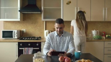 Mutlu genç adam kız arkadaşını evde yemek yaparken mutfakta oturmuş dijital tablet bilgisayar kullanarak