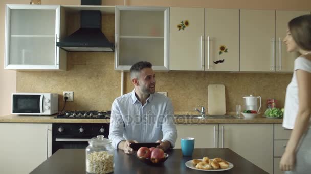 Atraente jovem casal abraçando e beijando de manhã cedo na cozinha, enquanto o homem toma café da manhã — Vídeo de Stock