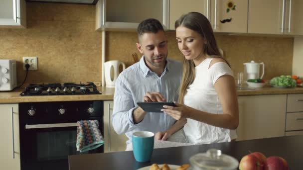 Счастливая молодая пара пользуется цифровым планшетным компьютером, сидя на кухне и завтракая утром — стоковое видео