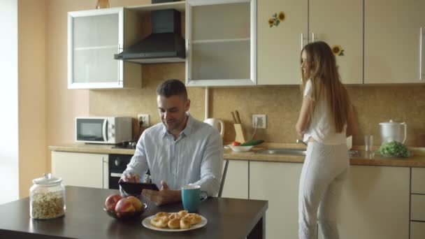 Jovem feliz usando computador tablet digital sentado na cozinha e falando com sua esposa enquanto ela cozinha em casa — Vídeo de Stock