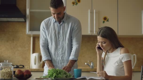 Casal atraente na cozinha de manhã cedo. Menina bonita conversando telefone enquanto seu namorado cozinhar café da manhã — Vídeo de Stock