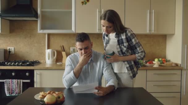 Uspet giovane uomo a leggere le bollette non pagate e abbracciato dalla moglie che lo sostiene in cucina a casa — Video Stock