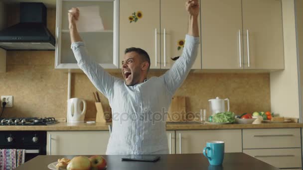 Hombre guapo recibir buenas noticias carta de lectura en la cocina mientras desayuna en casa temprano por la mañana — Vídeo de stock