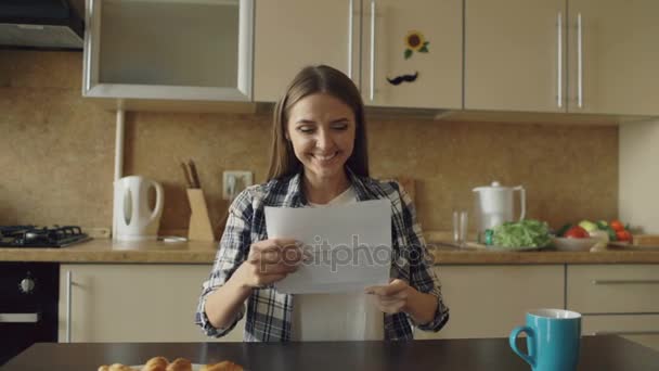 Attraktive, glückliche Frau erhält gute Nachrichten beim Lesen von Briefen in der Küche, während sie frühmorgens zu Hause frühstückt — Stockvideo