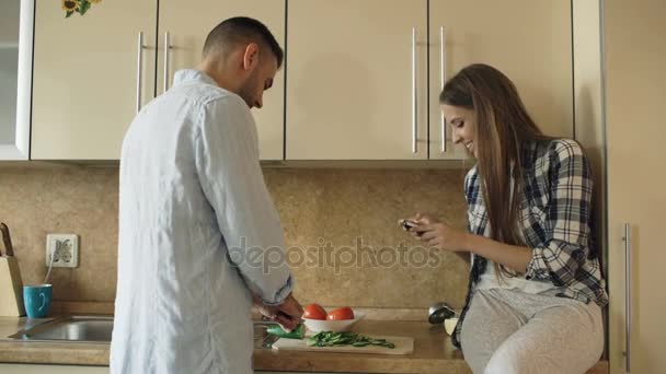 Casal atraente cozinhar na cozinha e tirar fotos usando o smartphone fo compartilhamento de mídia social em casa — Vídeo de Stock