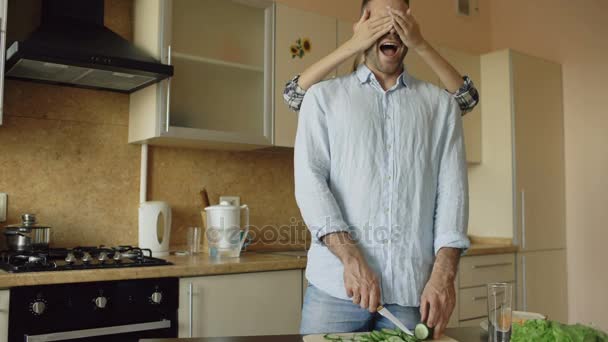 Молодая женщина закрывает парням глаза руками и удивляет его на кухне дома — стоковое видео