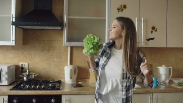 Vrolijke jonge grappige vrouw dansen en zingen met sla microfoon tijdens het ontbijt koken — Stockvideo