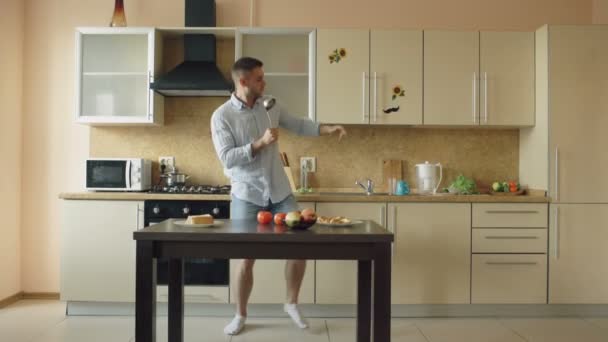 Привлекательный молодой смешной человек танцует и поет с ковш во время приготовления пищи на кухне дома — стоковое видео