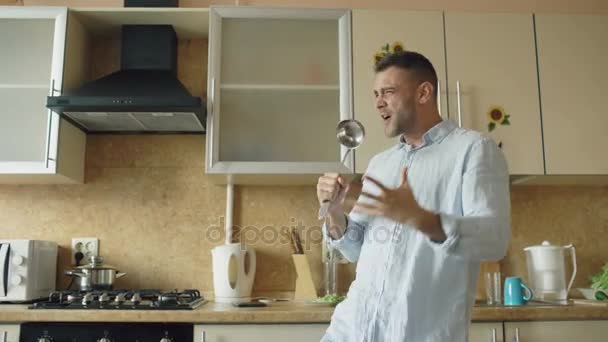 Aantrekkelijke jonge grappige man dansen en zingen met pollepel tijdens het koken in de keuken thuis — Stockvideo