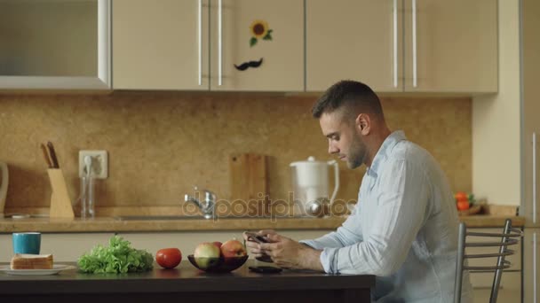 ながら台所に座ってデジタル タブレット コンピューターを使用して深刻な若い男がある朝の朝食 — ストック動画