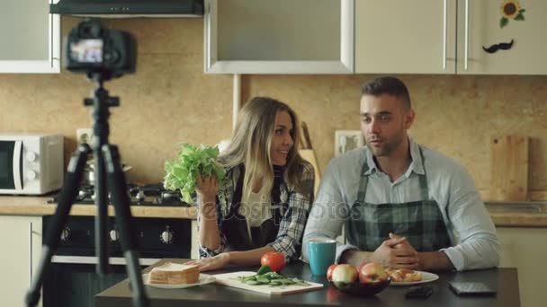 Jonge aantrekkelijke (echt) paar bloggers schieten video voedsel blog over koken op dslr camera in de keuken — Stockvideo