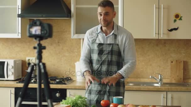 Jonge knappe man in schort schieten video voedsel blog over koken op dslr camera in keuken — Stockvideo