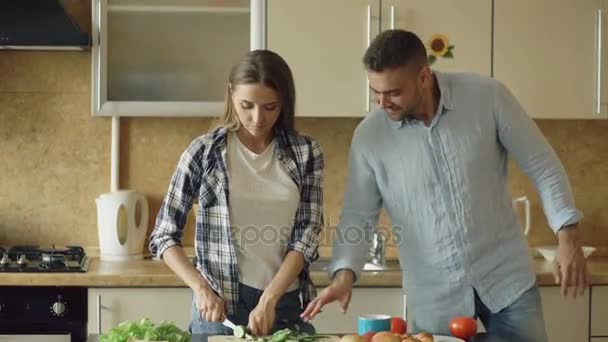 Um jovem casal feliz na cozinha. Mulher atraente cozinhar café da manhã e algema seu namorado que quer saborear comida — Vídeo de Stock