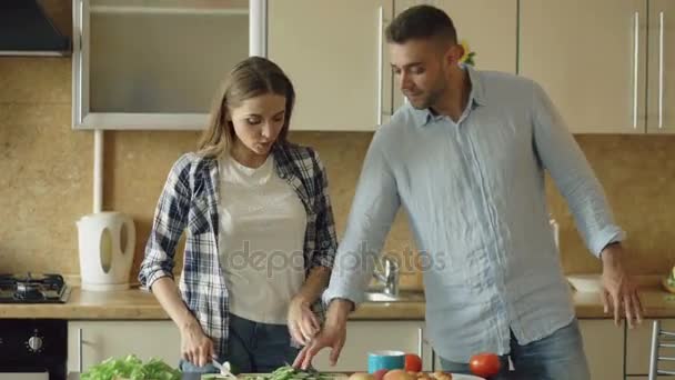 Boldog, fiatal pár, a konyhában. Vonzó nő főzés reggeli, és a barátja, aki akar az élelmiszer íze mandzsetta — Stock videók