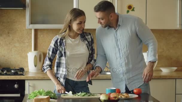 Boldog, fiatal pár, a konyhában. Vonzó nő főzés reggeli, és a barátja, aki akar az élelmiszer íze mandzsetta — Stock videók