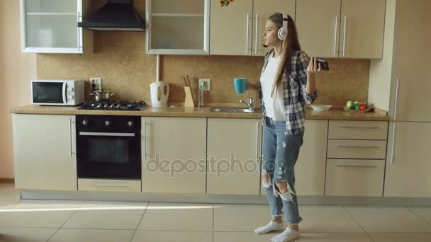 Attraktive junge lustige Frau mit Kopfhörer, die morgens in der Küche tanzt und singt und Spaß im Urlaub hat — Stockvideo