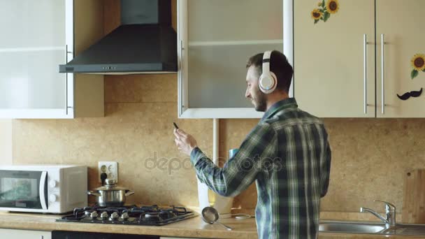 Bonito jovem engraçado homem em fones de ouvido dançando e cantando na cozinha em casa de manhã e se divertir em férias — Vídeo de Stock