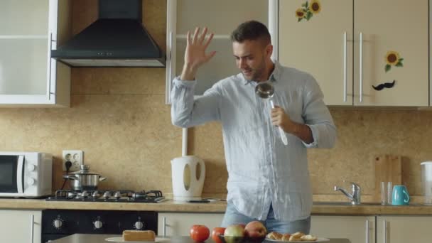 Slow motion van aantrekkelijke jonge grappige man dansen en zingen met pollepel tijdens het koken in de keuken thuis — Stockvideo