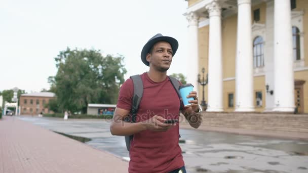 Steadicam disparo de estudiante sonriente en el sombrero caminando y surf teléfono inteligente beber café al aire libre — Vídeo de stock