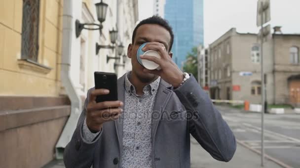 Steadicam shot of young happy businessman using smartphone and walking with cup of coffee outdoors — Stock Video