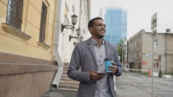 Steadicam plan de jeune homme d'affaires heureux en utilisant smartphone et marcher avec une tasse de café à l'extérieur — Video