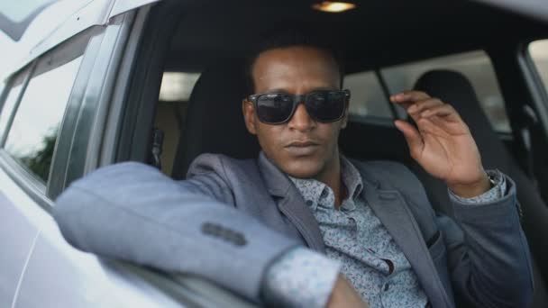 Портрет серьезного бизнесмена, сидящего в машине, надевающего солнцезащитные очки и улыбающегося в камеру на улице — стоковое видео